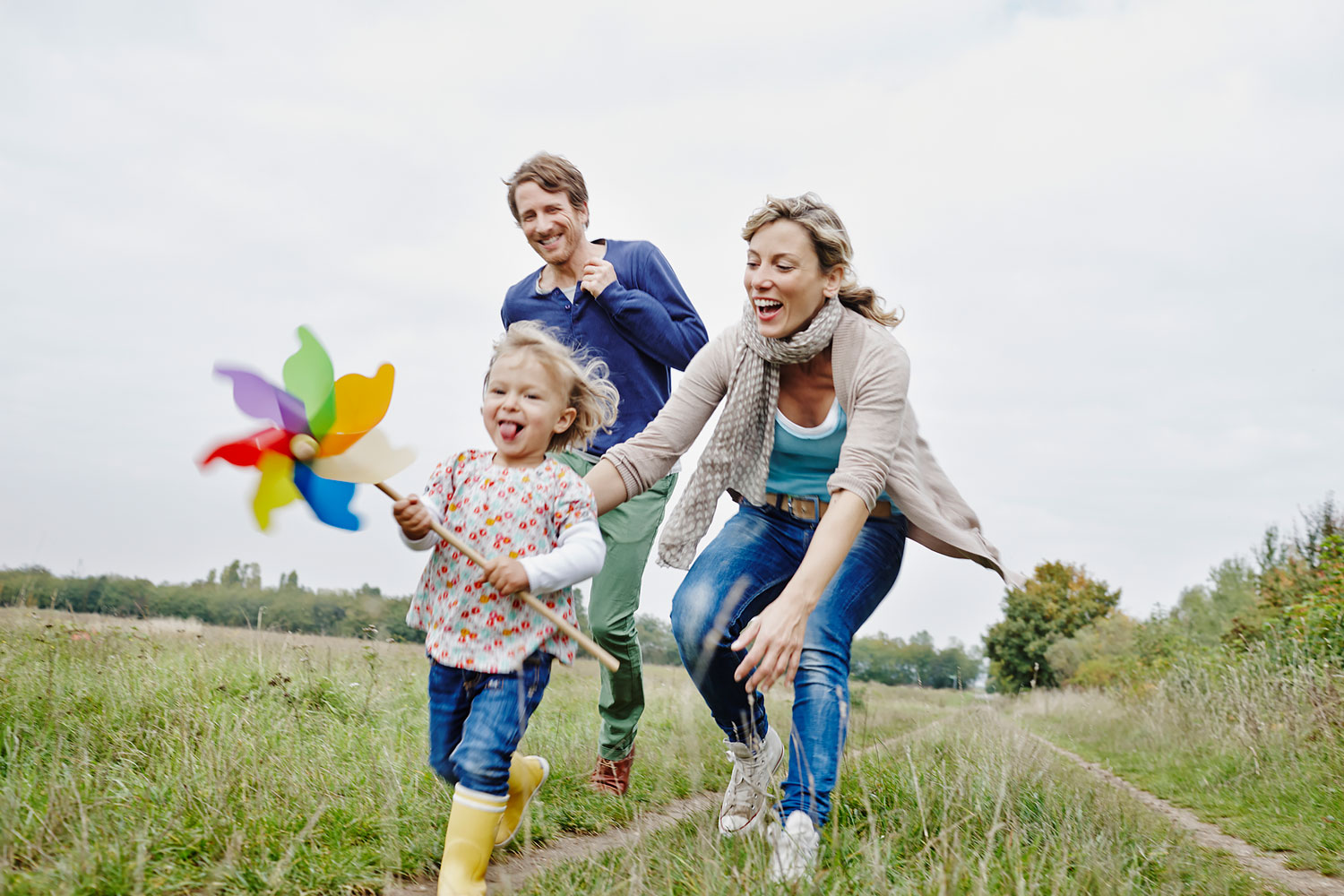A family—mother, father, and daughter—is running across a meadow, laughing; the daughter is holding a colorful pinwheel in her hand