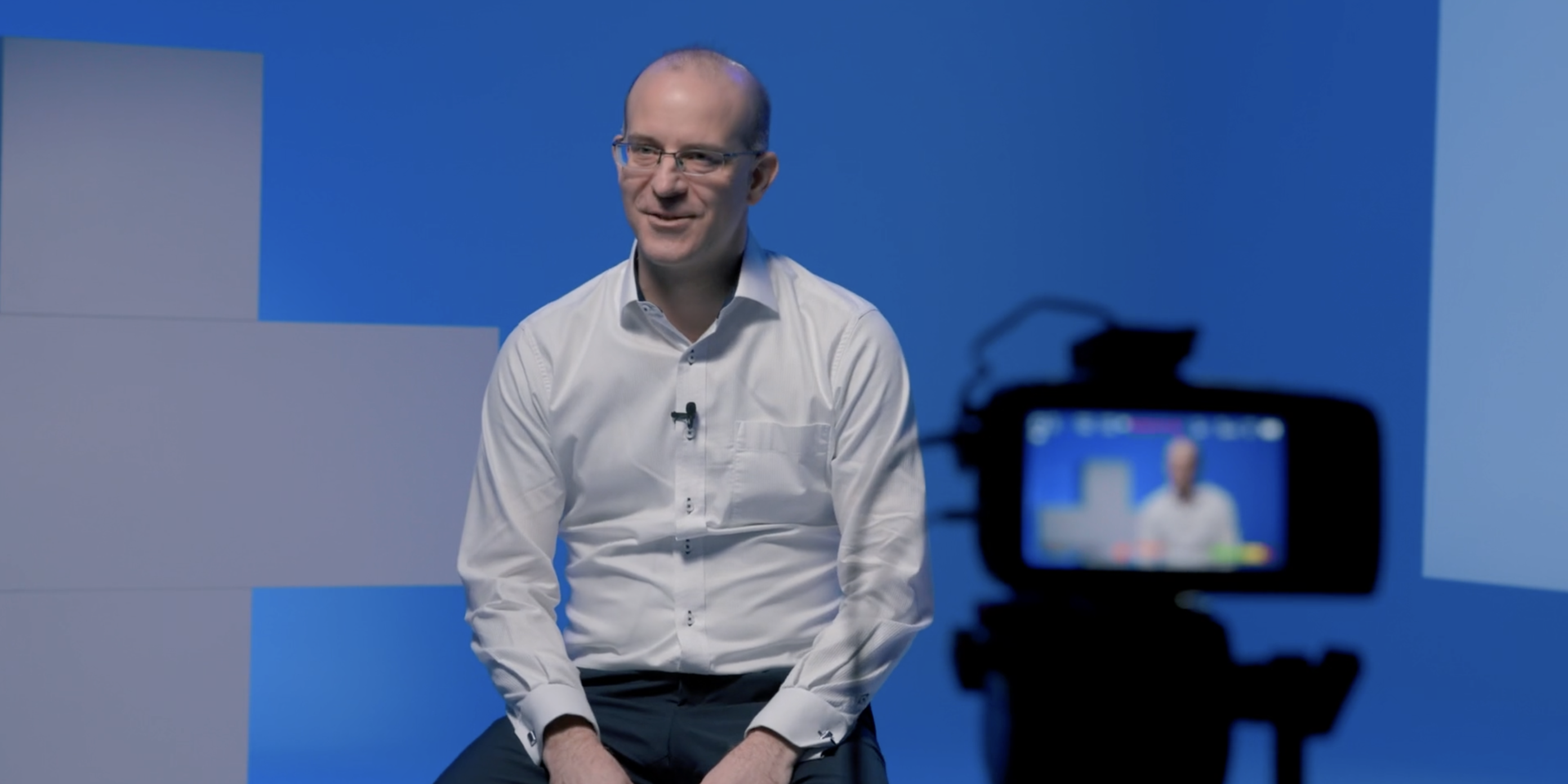 Our CSO Erik Edelmann is wearing a white shirt and sitting behind a camera. He is currently being interviewed and the interview is being filmed