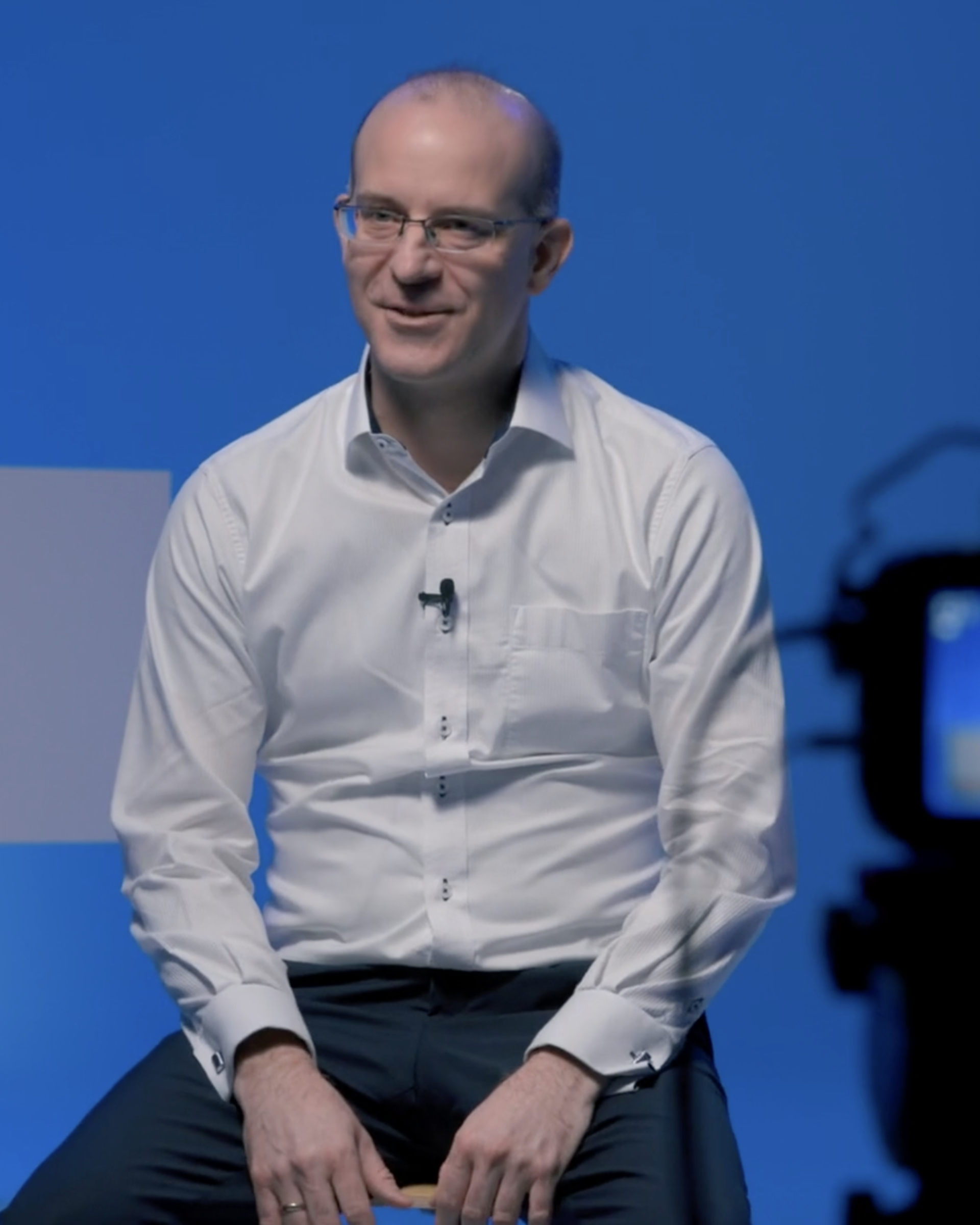 Unser CSO Erik Edelmann trägt ein weißes Hemd und sitzt hinter einer Kamera. Er wird gerade interviewt und das Interview gefilmt
