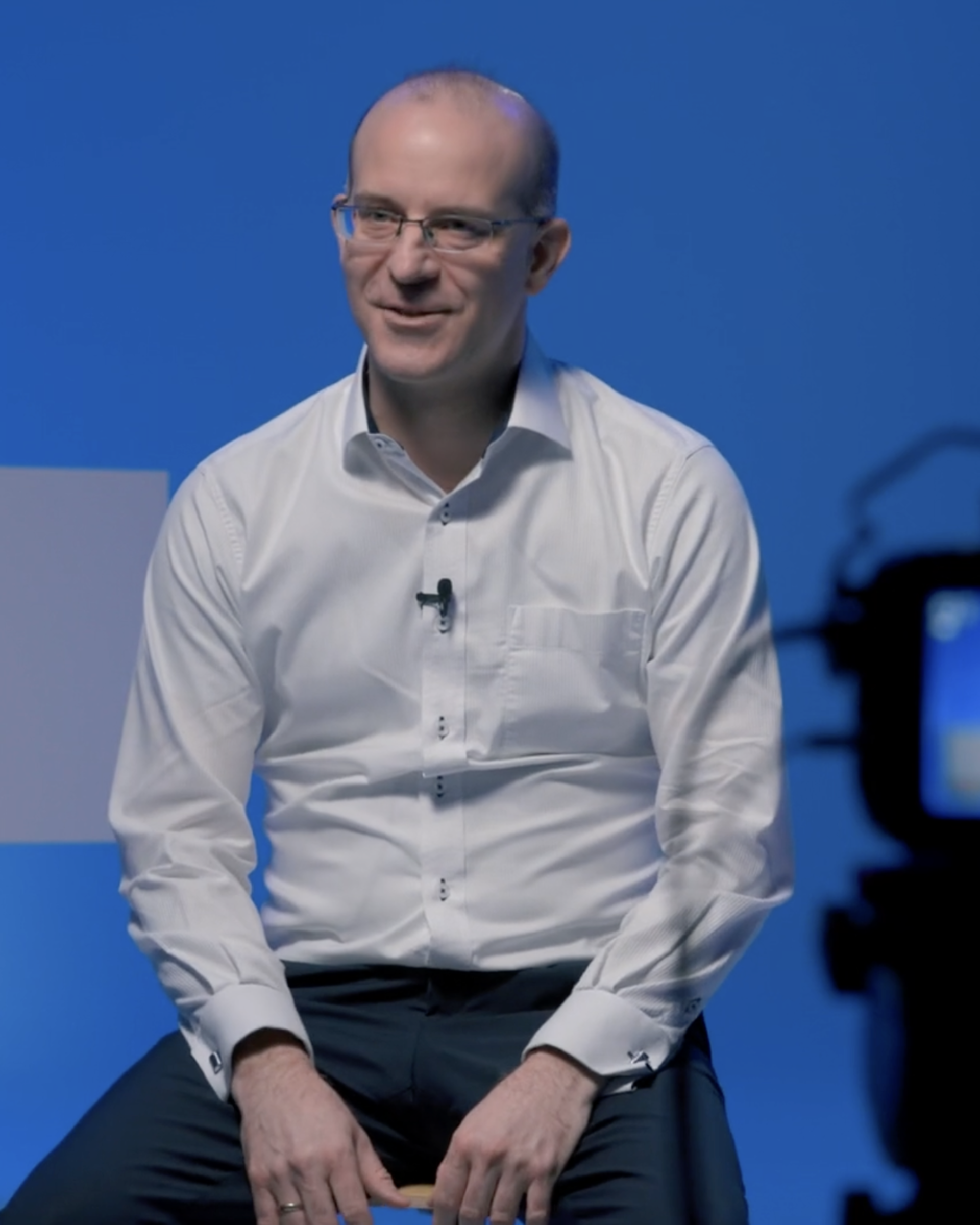 Our CSO Erik Edelmann is wearing a white shirt and sitting behind a camera. He is currently being interviewed and the interview is being filmed