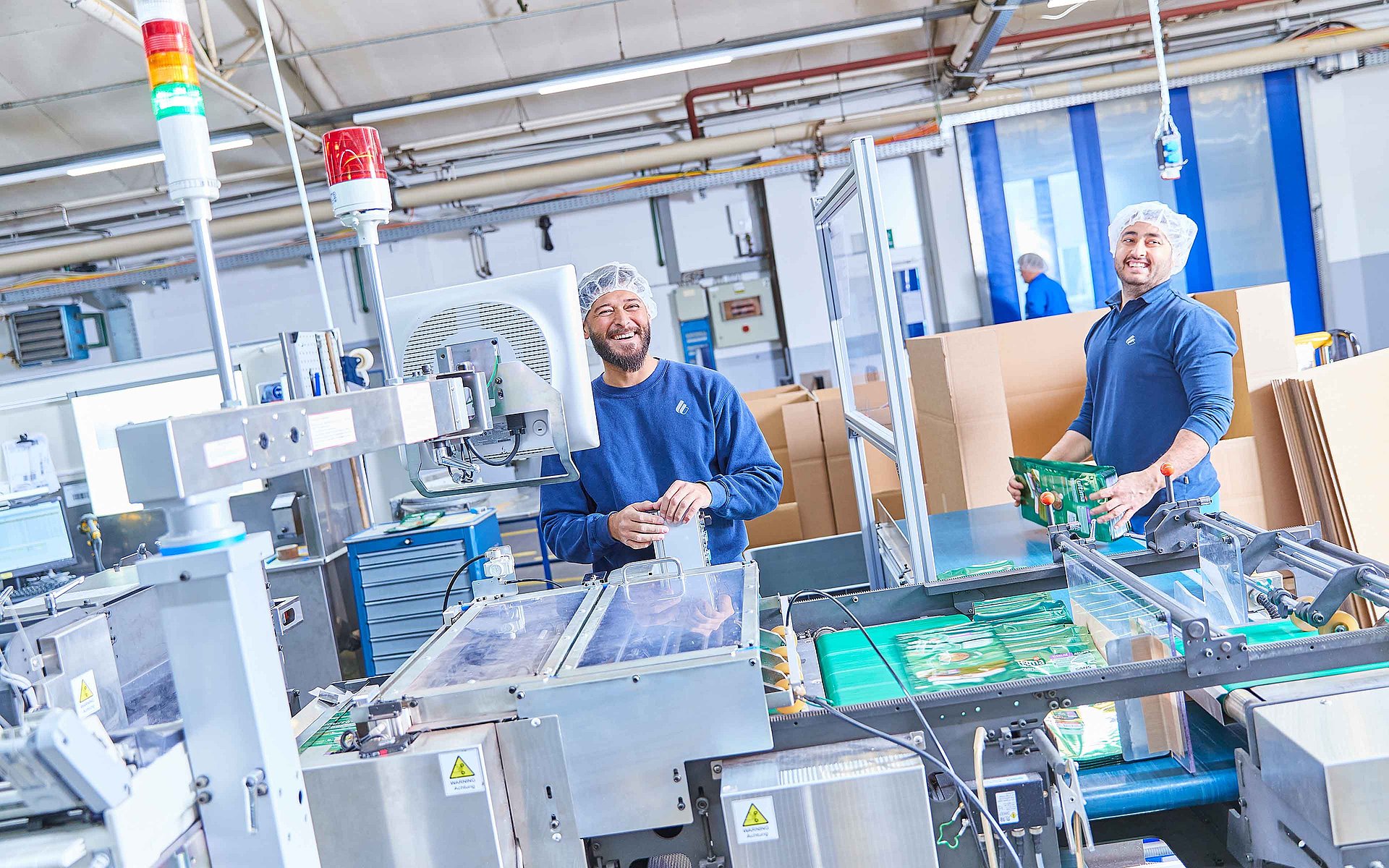 Dwóch pracowników w ubraniach roboczych stoi przy maszynie produkcyjnej i trzyma w rękach gotowe opakowania na paszę dla zwierząt. Uśmiechają się do kamery.