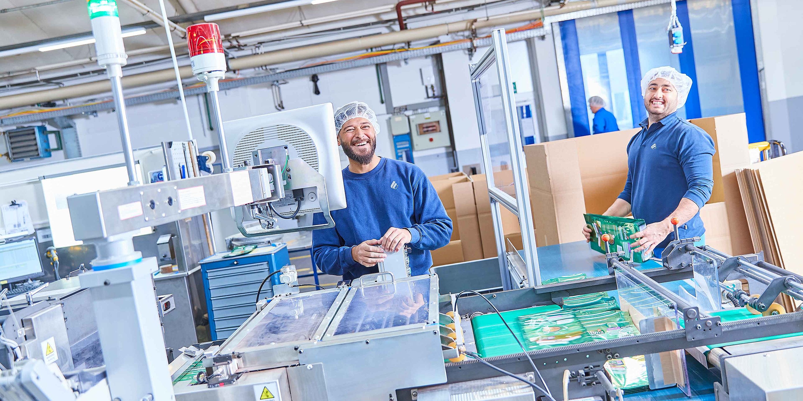 Two employees in work clothing are standing by a production machine and are holding finished pet food packaging in their hands. They are smiling into the camera.