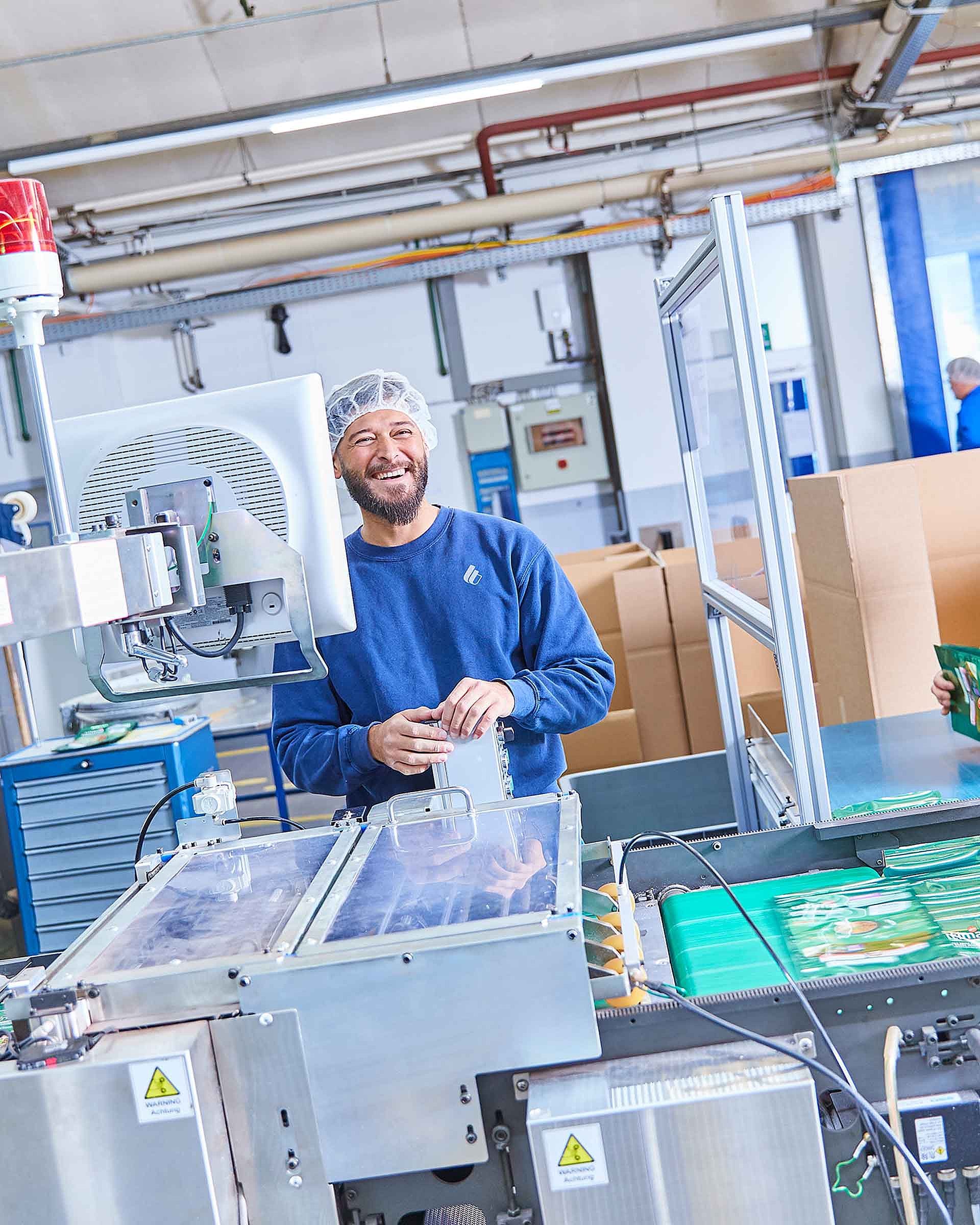 Two employees in work clothing are standing by a production machine and are holding finished pet food packaging in their hands. They are smiling into the camera.
