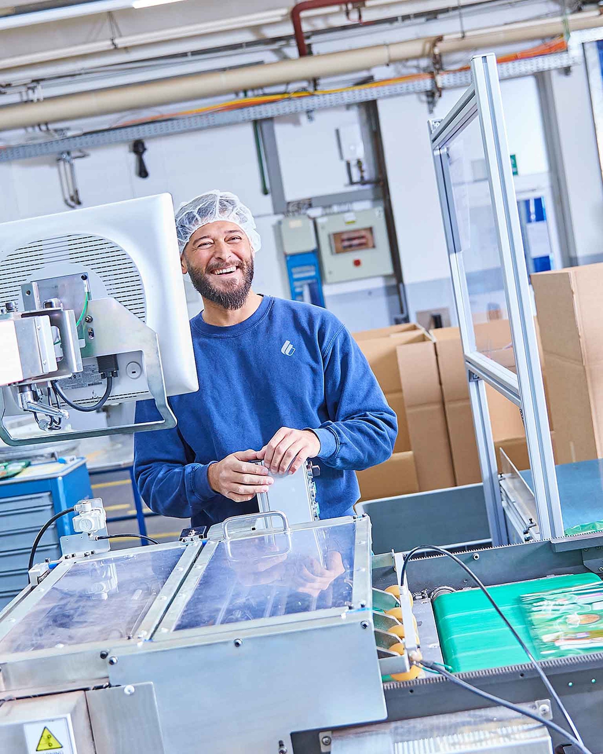 Zwei Mitarbeitende in Arbeitskleidung stehen an einer Produktionsmaschine und halten fertig produzierte Tierfutter-Verpackungen in der Hand. Sie lächeln in die Kamera.
