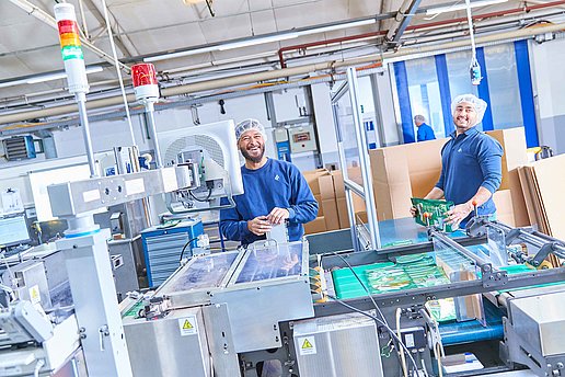 Two employees in work clothing are standing by a production machine and are holding finished pet food packaging in their hands. They are smiling into the camera.