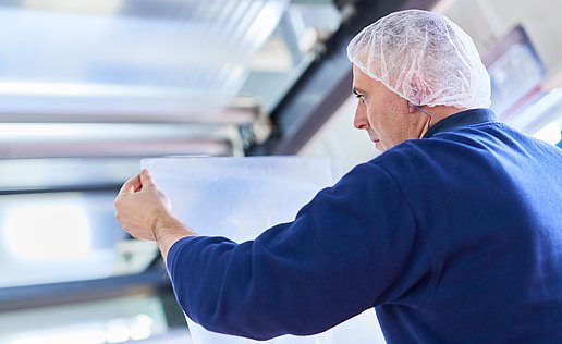 An employee in dark blue work clothing and a hair net can be seen from behind. He is holding a plastic film in his hands