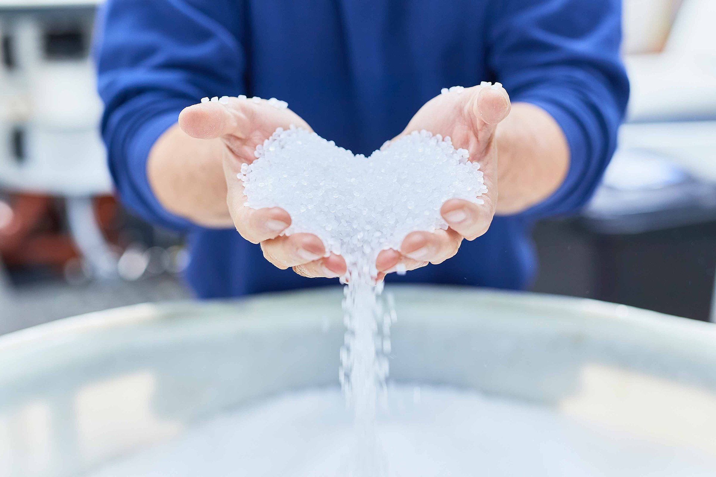 Two hands are pouring out white plastic granules in the shape of a heart. They are used to produce flexible plastic packaging