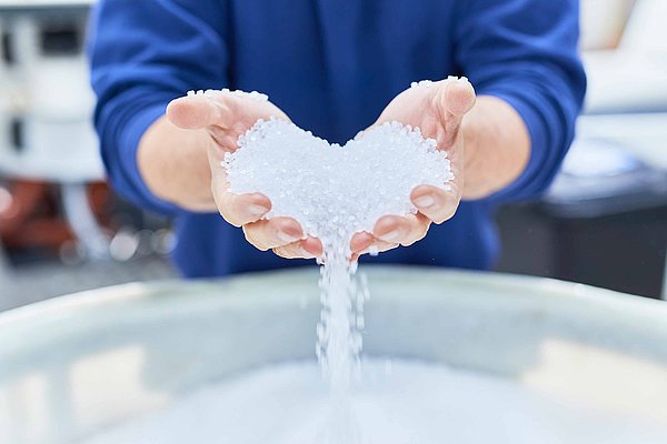 Two hands are pouring out white plastic granules in the shape of a heart. They are used to produce flexible plastic packaging