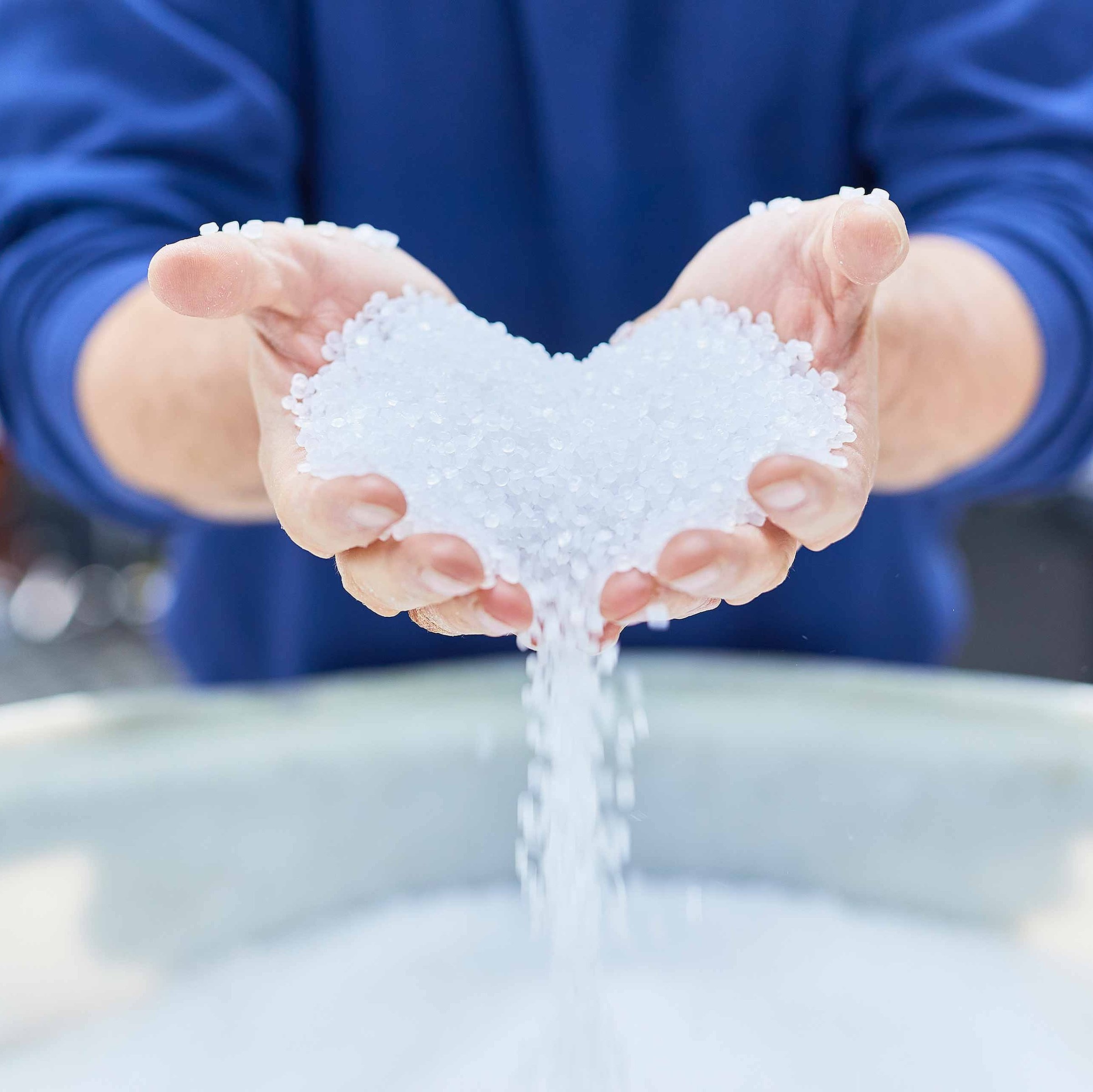 Two hands are pouring out white plastic granules in the shape of a heart. They are used to produce flexible plastic packaging