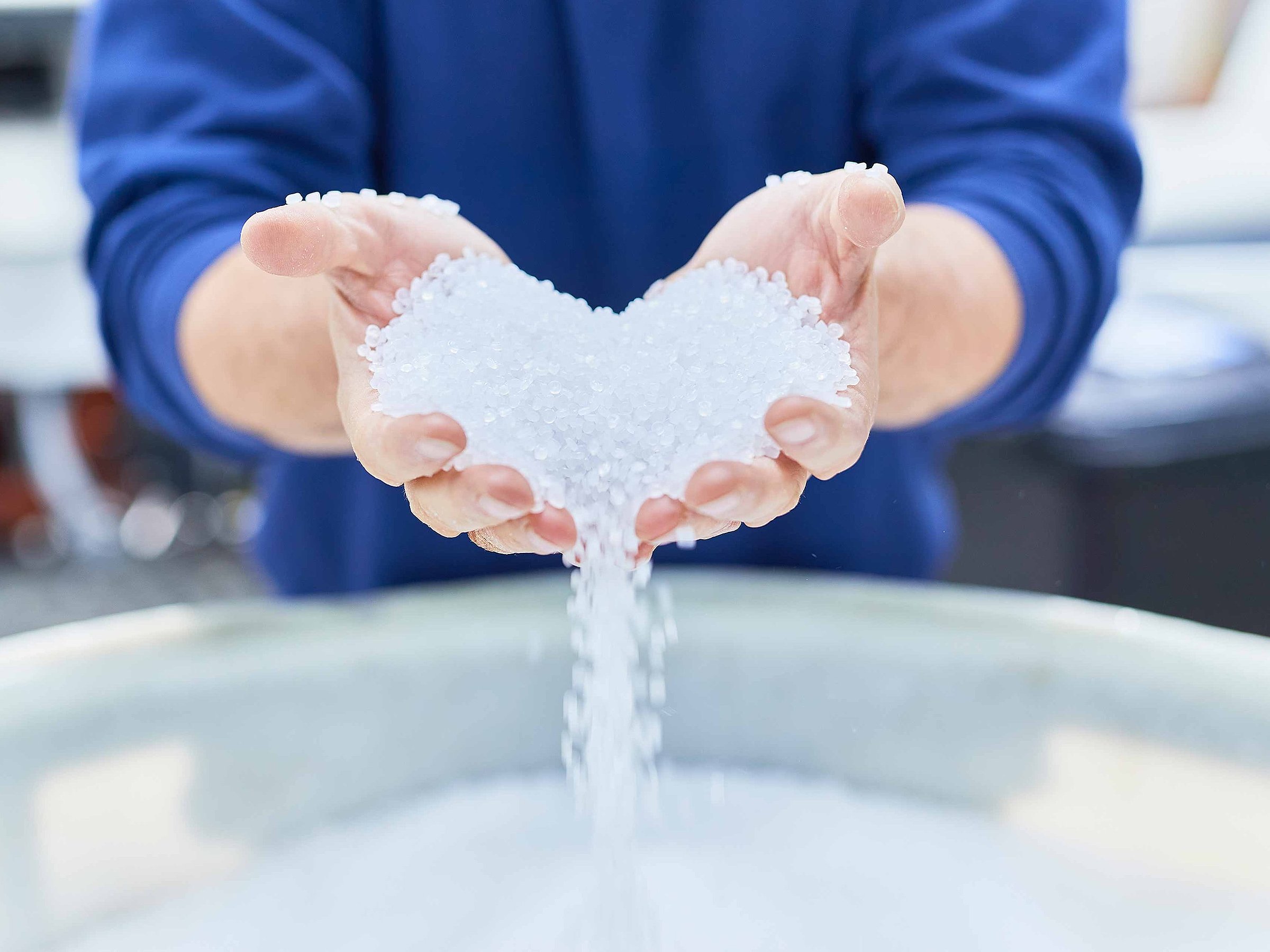 Two hands are pouring out white plastic granules in the shape of a heart. They are used to produce flexible plastic packaging