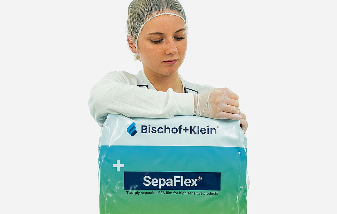 Une collaboratrice porte des vêtements de travail blancs, une résille et des gants. Elle tient un emballage plastique dans sa main et veut l'ouvrir. 