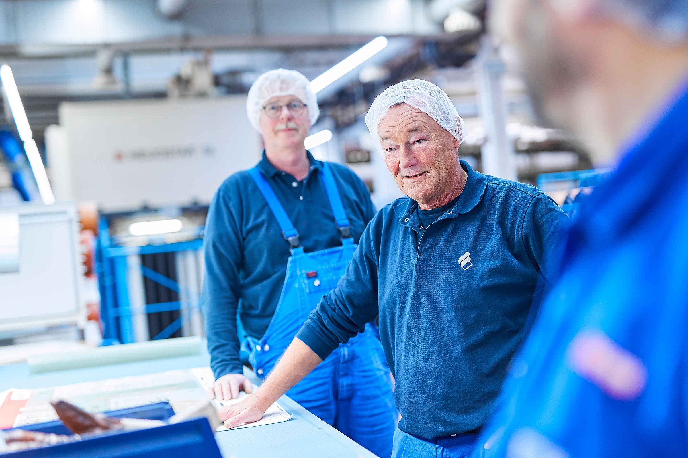 The picture shows two Bischof+Klein employees working in the production department. Both are wearing blue work clothing. The picture is focused on one employee who is explaining something. The other employee is standing more in the background