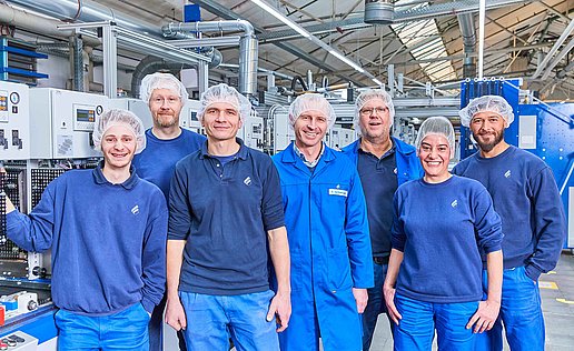 The picture shows seven Bischof+Klein employees. They are all wearing dark blue work clothing and hair nets and are standing in the production hall
