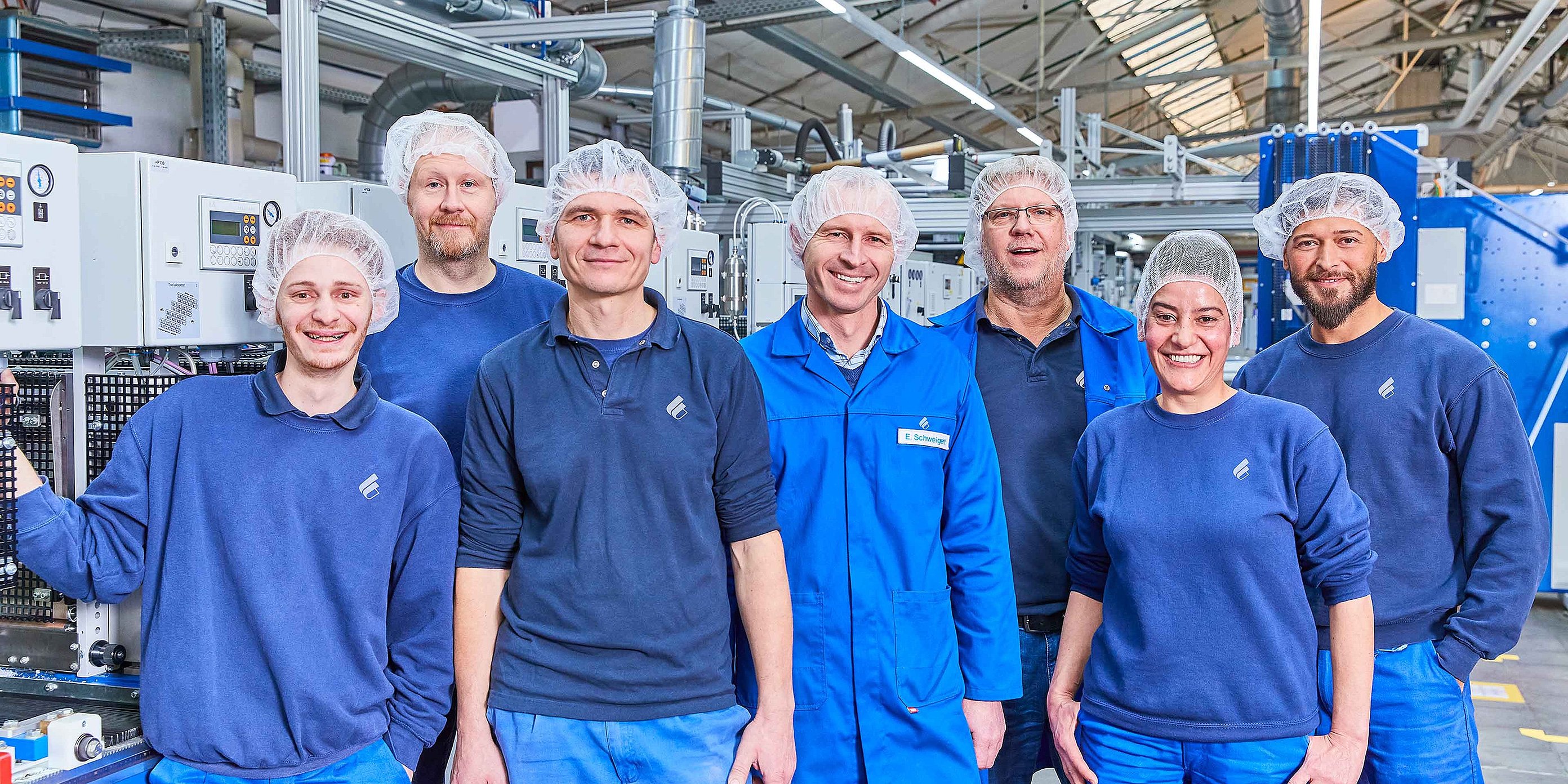 Zu sehen sind sieben Mitarbeitende von Bischof+Klein. Sie tragen alle dunkelblaue Arbeitskleidung und Haarnetze und stehen in der Produktionshalle