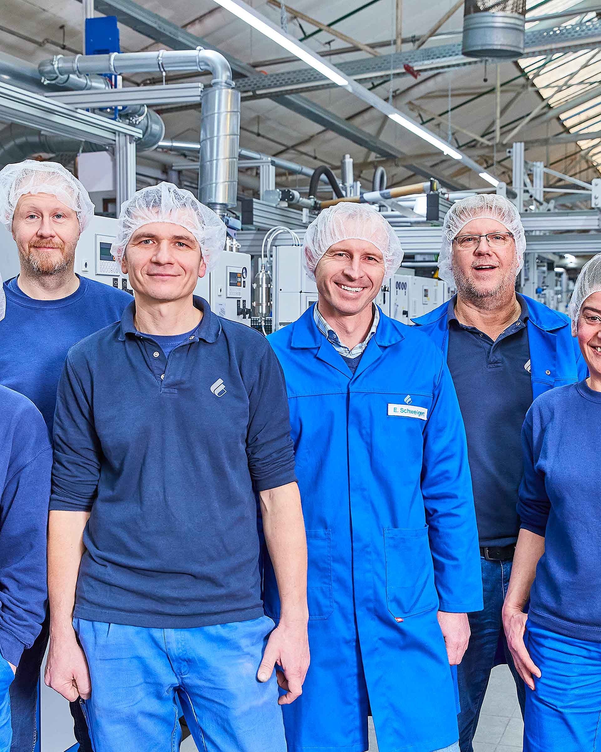 The picture shows seven Bischof+Klein employees. They are all wearing dark blue work clothing and hair nets and are standing in the production hall