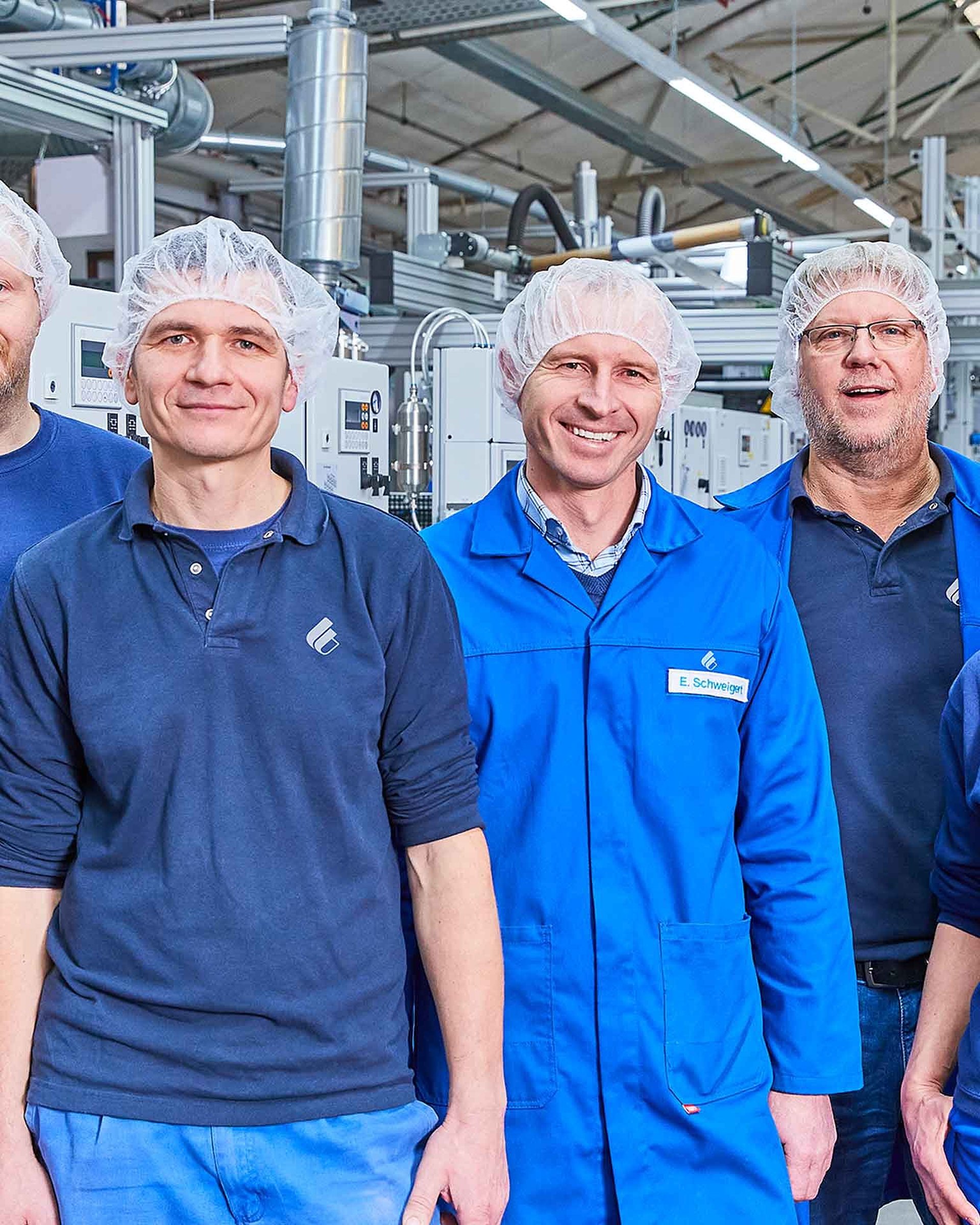 Zu sehen sind sieben Mitarbeitende von Bischof+Klein. Sie tragen alle dunkelblaue Arbeitskleidung und Haarnetze und stehen in der Produktionshalle
