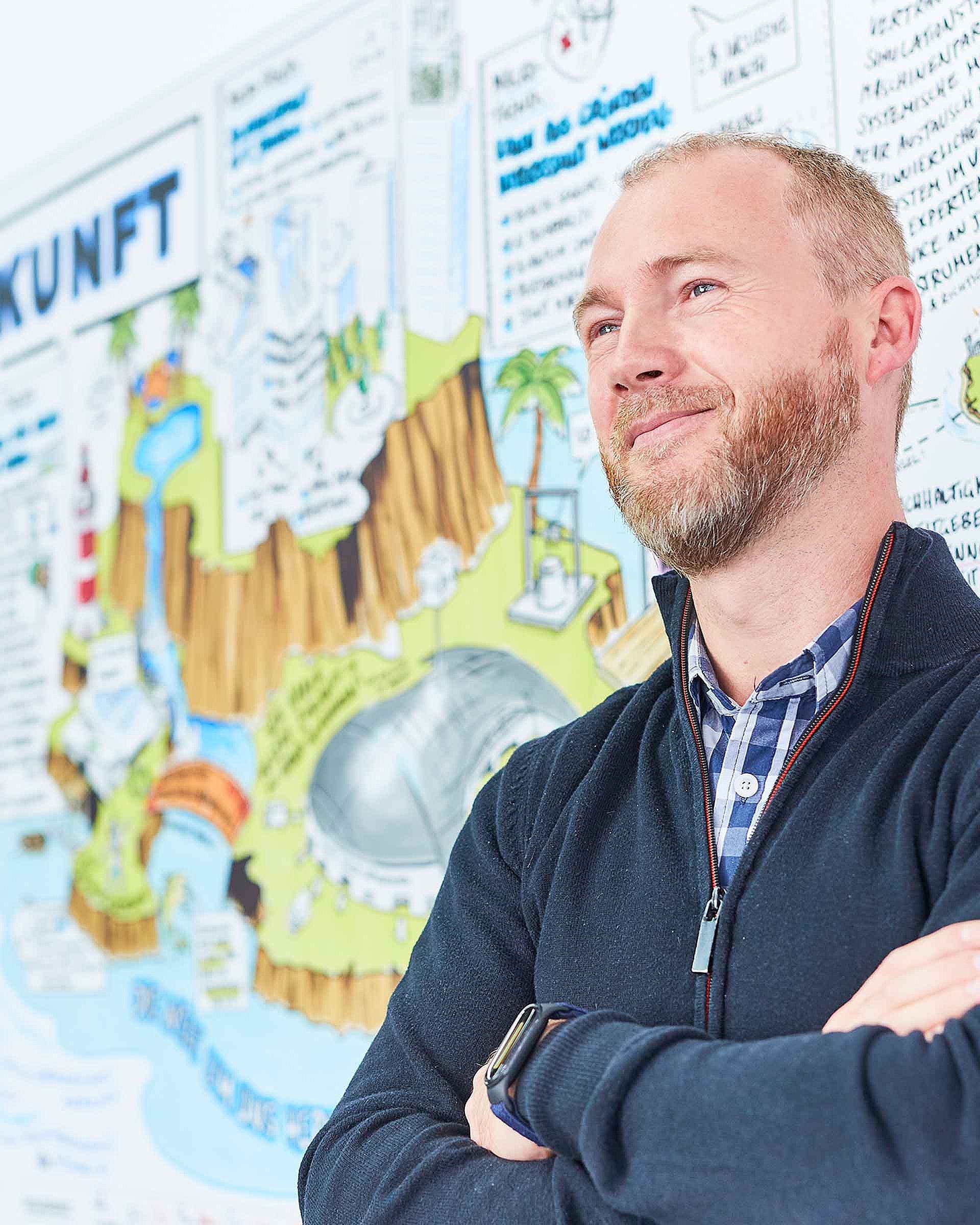 An employee is standing in front of a colorful wall. The values of Bischof+Klein can be seen painted as a blurred comic strip in the background.