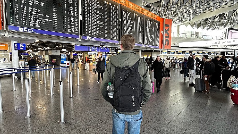 Der ehemalige duale Student Florenz steht am Flughafen und schaut auf eine Anzeigetafel, auf der die Flüge aufgelistet sind