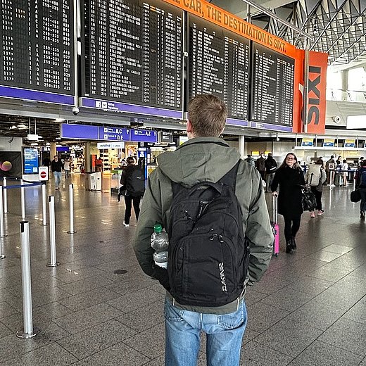 Der ehemalige duale Student Florenz steht am Flughafen und schaut auf eine Anzeigetafel, auf der die Flüge aufgelistet sind