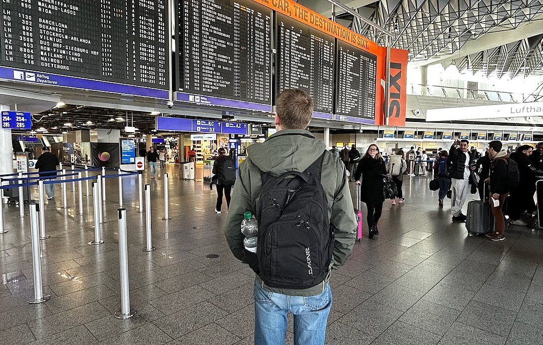 Der ehemalige duale Student Florenz steht am Flughafen und schaut auf eine Anzeigetafel, auf der die Flüge aufgelistet sind