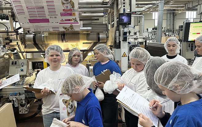 Mehrere Schulkinder tragen weiße Schutzmäntel und stehen in der Produktion an einer Produktionsanlage. Sie halten ein Klemmbrett in der Hand und notieren etwas