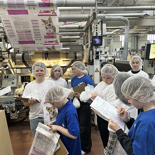 Mehrere Schulkinder tragen weiße Schutzmäntel und stehen in der Produktion an einer Produktionsanlage. Sie halten ein Klemmbrett in der Hand und notieren etwas