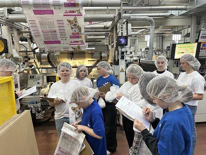 Mehrere Schulkinder tragen weiße Schutzmäntel und stehen in der Produktion an einer Produktionsanlage. Sie halten ein Klemmbrett in der Hand und notieren etwas
