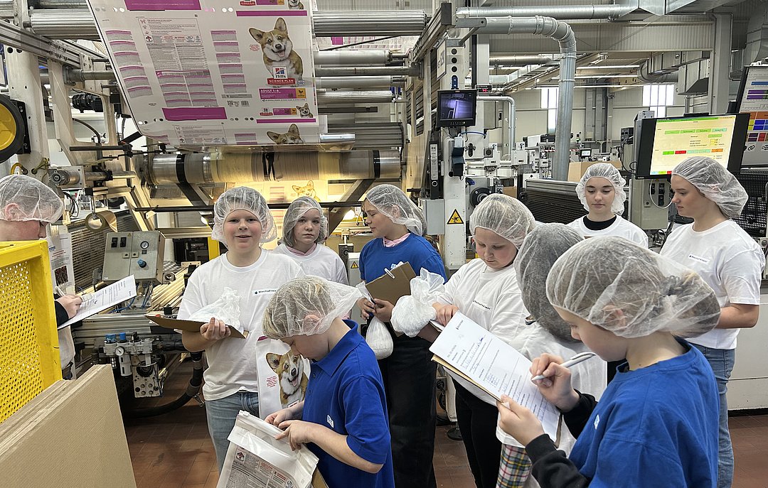 Mehrere Schulkinder tragen weiße Schutzmäntel und stehen in der Produktion an einer Produktionsanlage. Sie halten ein Klemmbrett in der Hand und notieren etwas