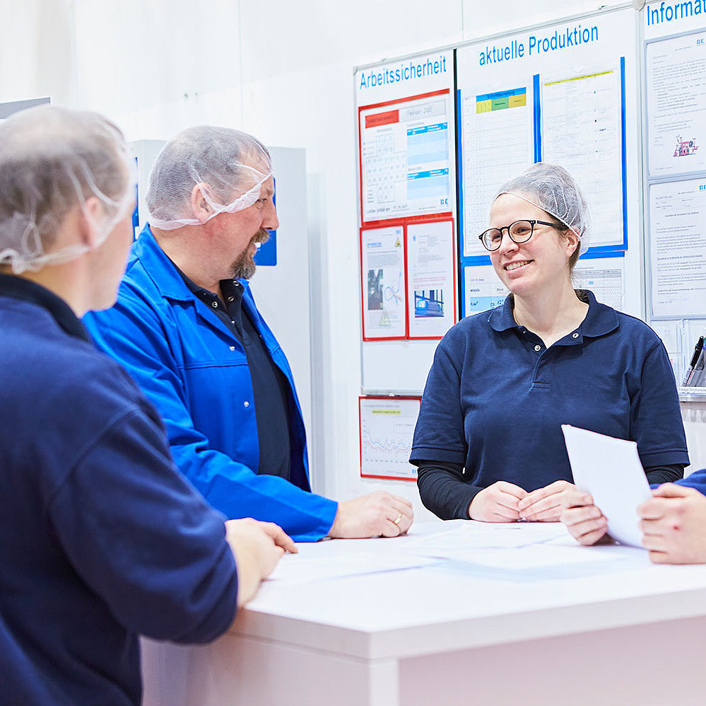 Three employees in work clothes are standing around a meeting table in the production area, chatting. In the background is a magnetic bulletin board displaying information on workplace safety and production.