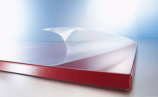Eine durchsichtige TacFlex® Oberflächenschutzfolien für Möbeloberflächen wird von einer roten Möbelfläche abgezogen