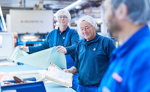 The picture shows two Bischof+Klein employees working in the production department. Both are wearing blue work clothing. The picture is focused on one employee who is explaining something. The other employee is standing more in the background. They are both holding plastic films in their hands