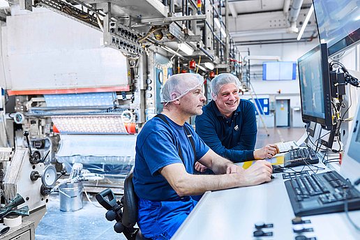 Two employees wearing Bischof+Klein work clothing are standing next to one another at a machine. They are wearing white hair nets and looking at a control panel on the machine