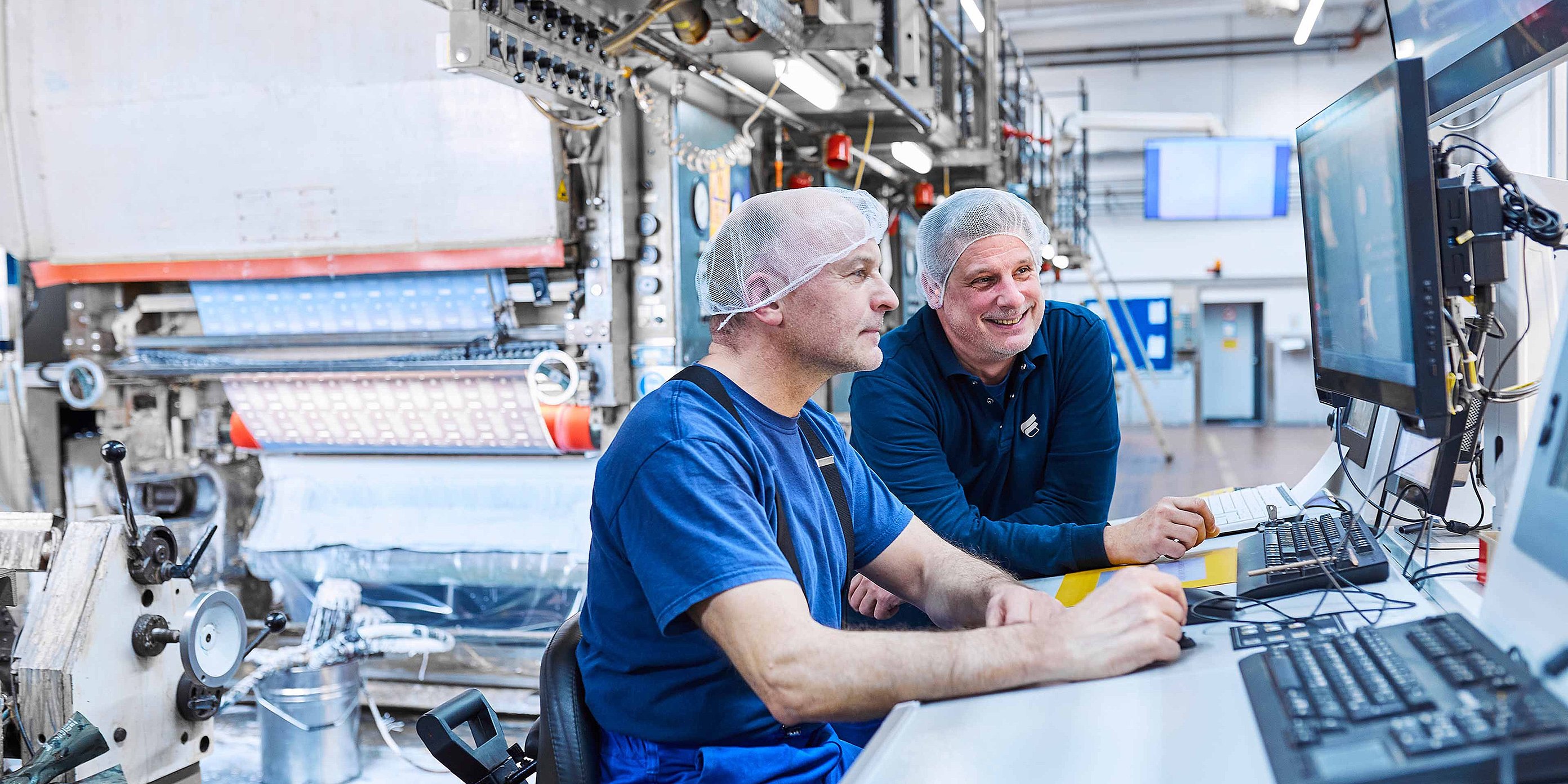 Zwei Mitarbeiter stehen in Bischof+Klein Arbeitskleidung nebeneinander an einer Maschine. Sie tragen weiße Haarnetze und schauen auf ein Bedienpanel der Maschine