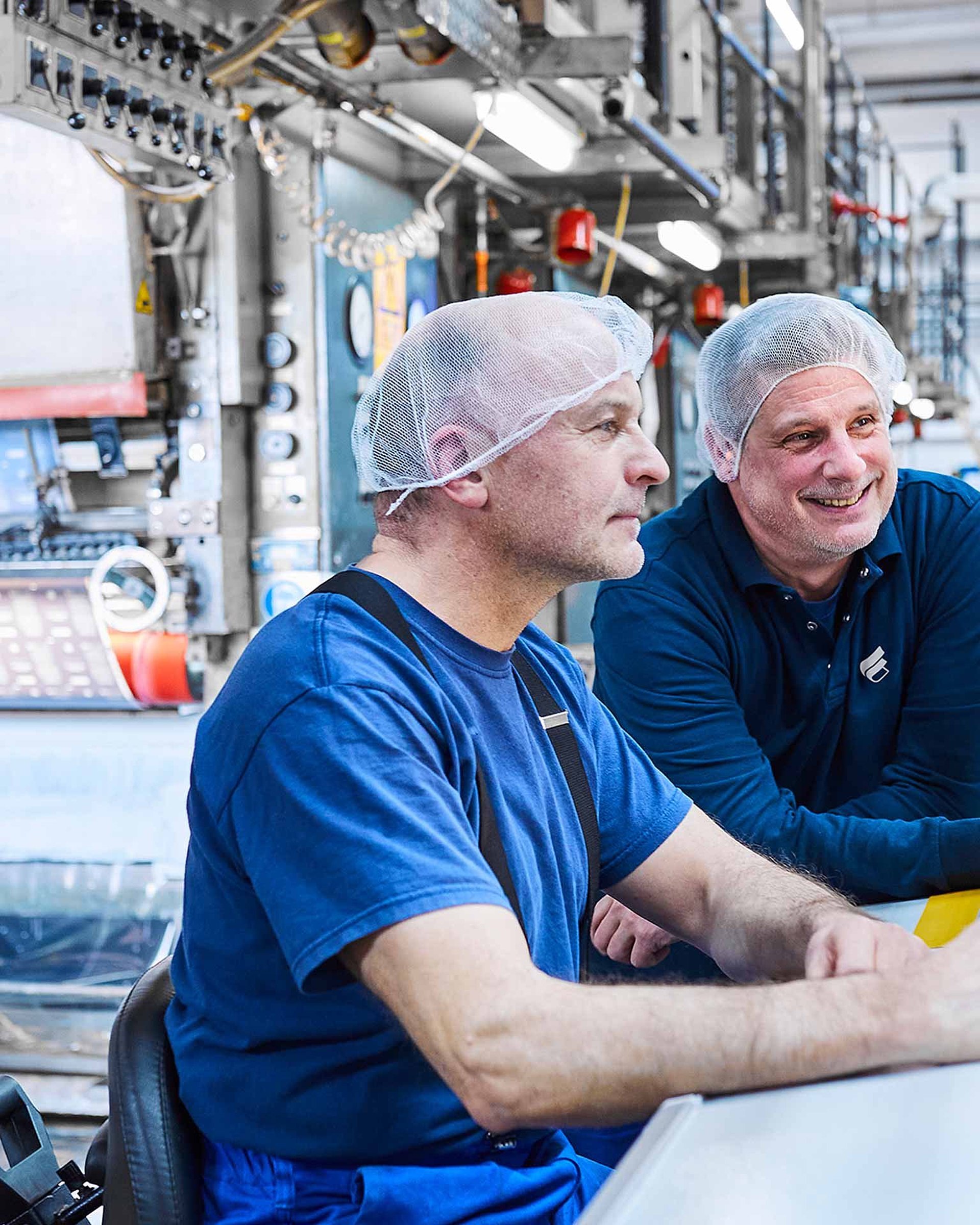 Zwei Mitarbeiter stehen in Bischof+Klein Arbeitskleidung nebeneinander an einer Maschine. Sie tragen weiße Haarnetze und schauen auf ein Bedienpanel der Maschine