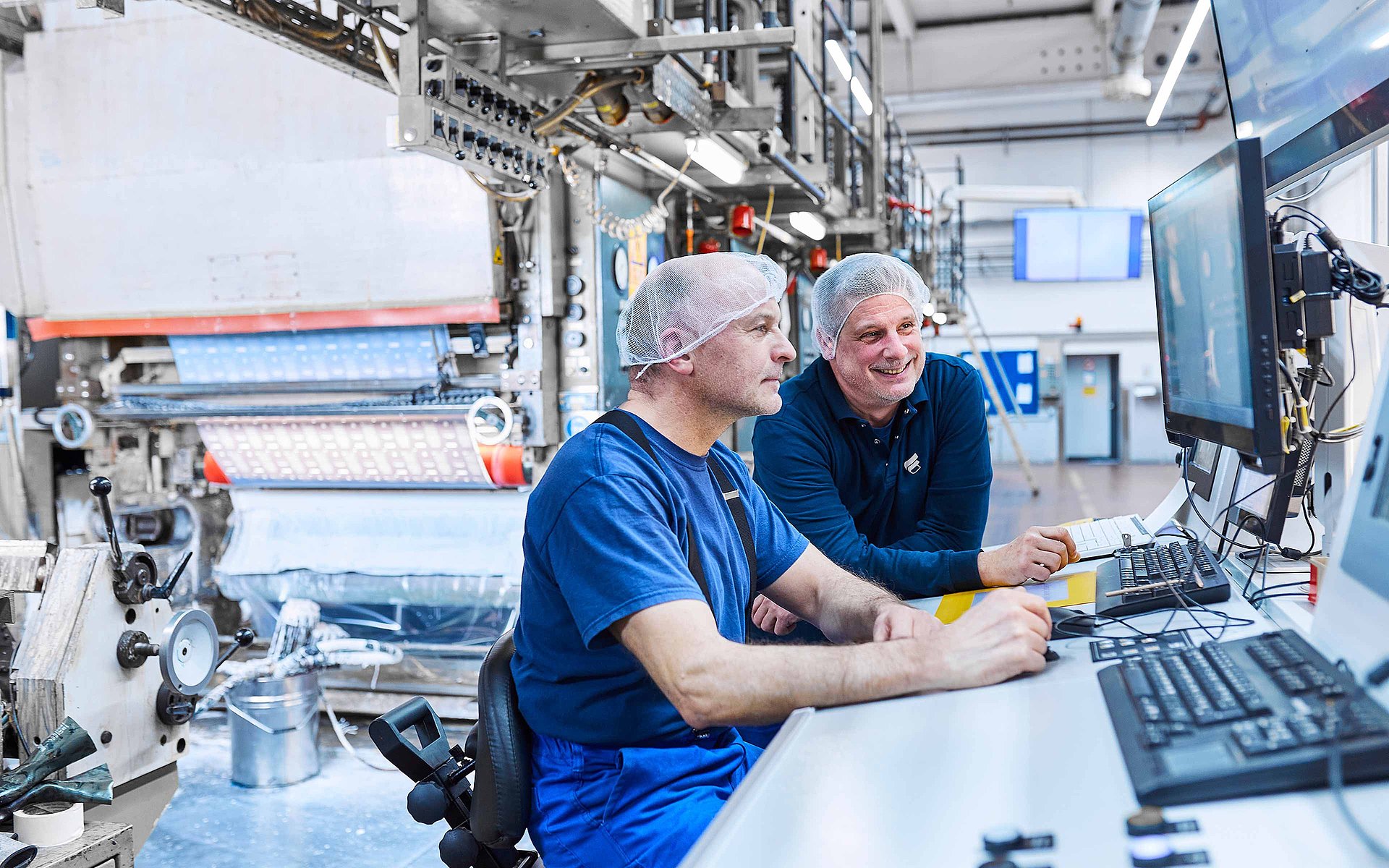 Deux collaborateurs en vêtement de travail de Bischof+Klein se tiennent côte à côte devant une machine. Ils portent des résilles blanches et regardent un pupitre de commande de la machine.