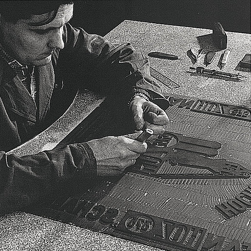 A man is working on a die cut with various tools at a table. It is a black and white photo 