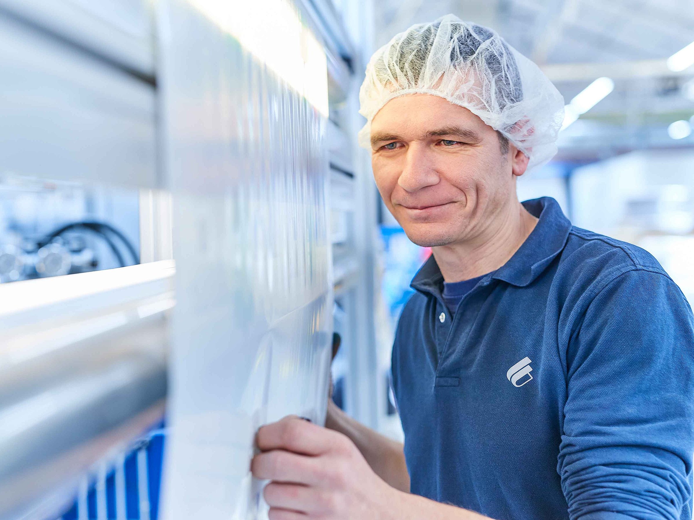 An employee wearing Bischof+Klein clothing and a hair net is standing behind a large reel of plastic film in the production department