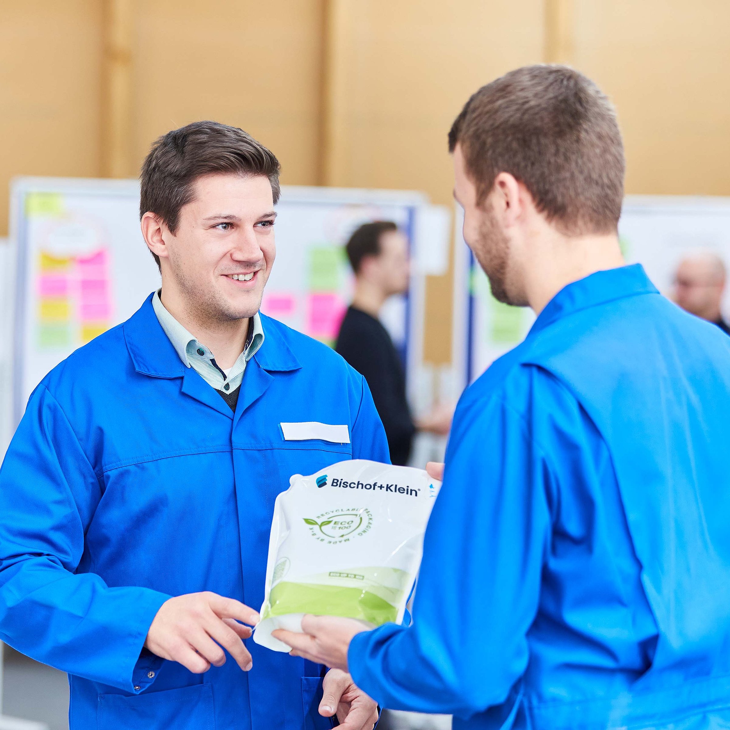 Two employees wearing work clothing are standing opposite one another and are discussing a product. One employee is holding a stand up pouch in his hand