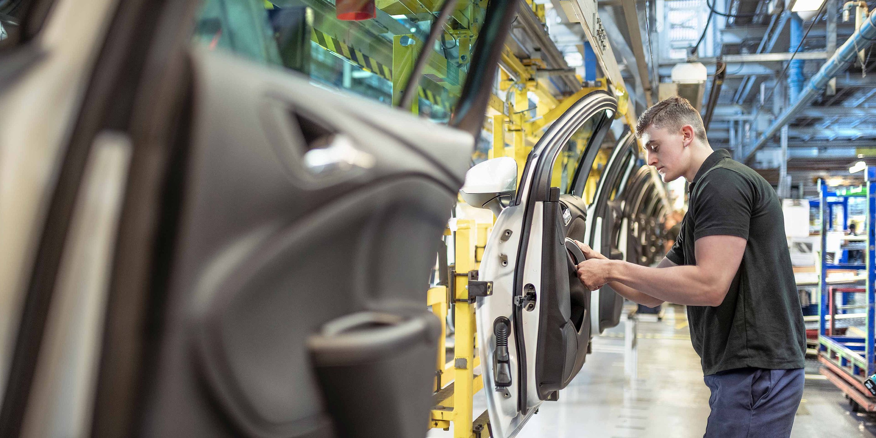 The picture shows a production hall in which cars are produced. A man is standing next to a car door fitting a part. He is wearing a black T-shirt