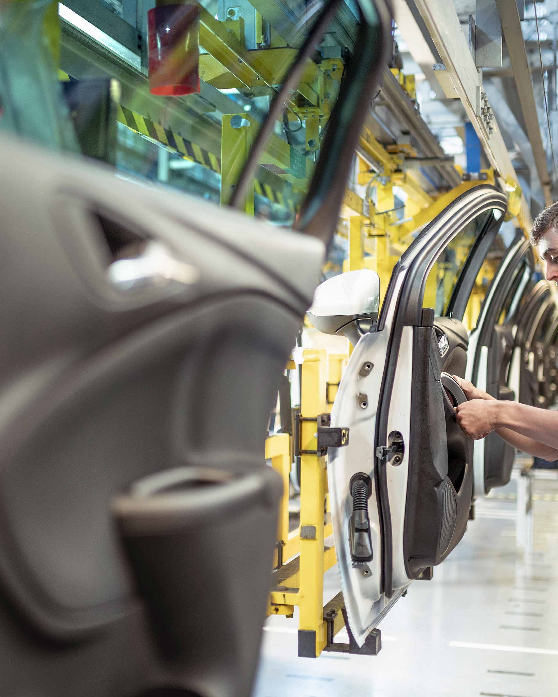 The picture shows a production hall in which cars are produced. A man is standing next to a car door fitting a part. He is wearing a black T-shirt