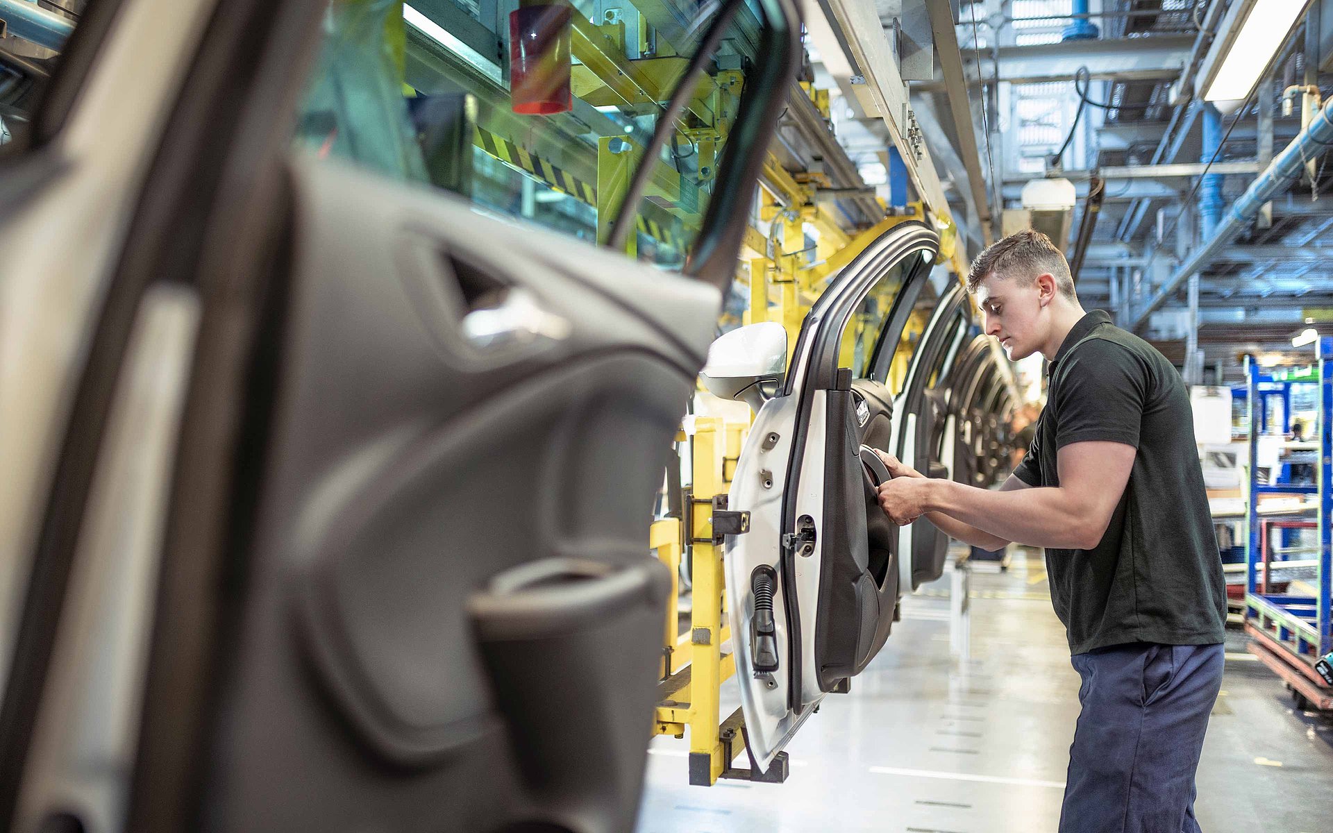 Un hall de production dans lequel des voitures sont fabriquées. Un homme se tient à côté d'une portière de voiture et monte une pièce. Il porte un t-shirt noir