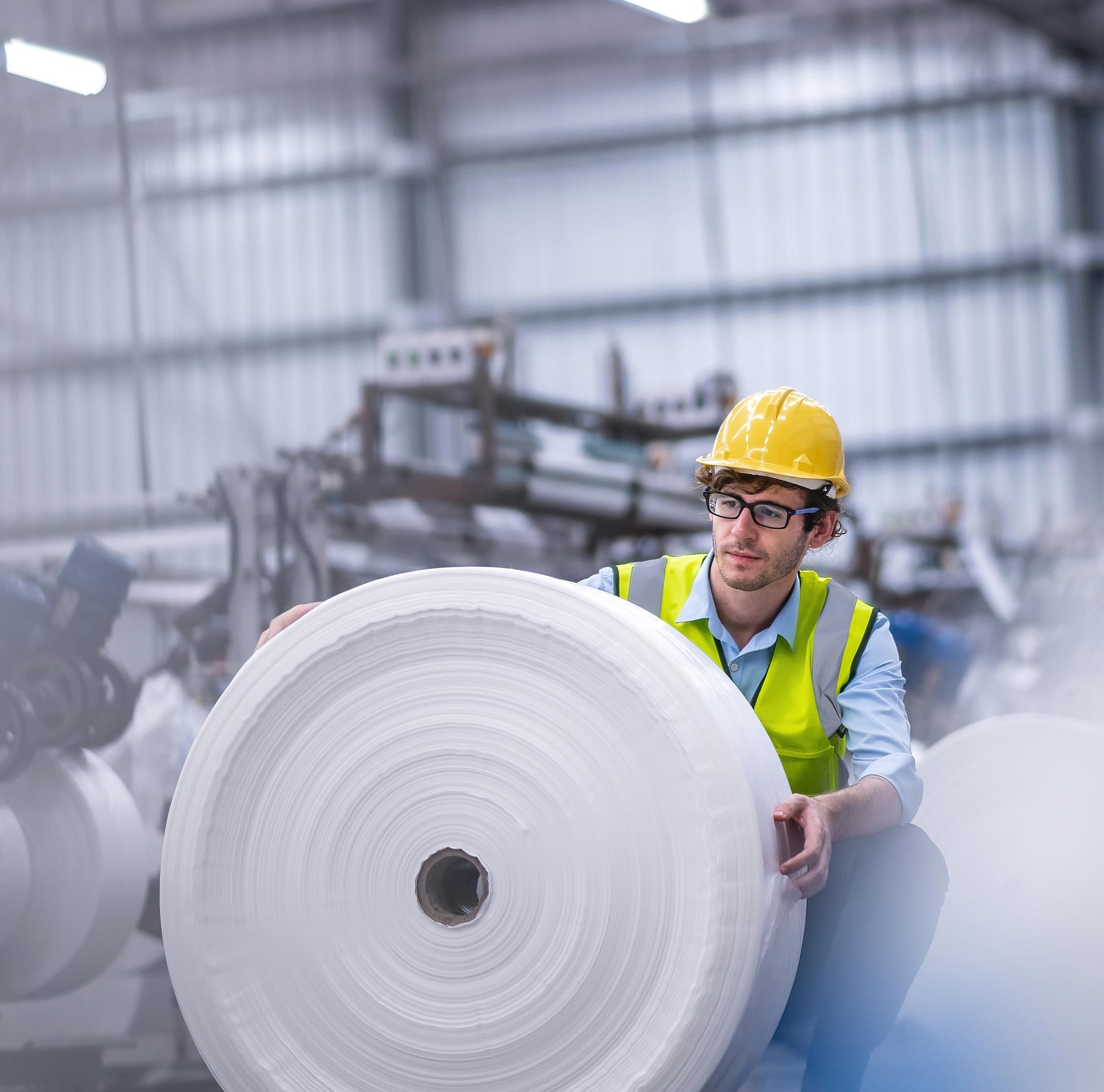 A man in a yellow hard hat and high-visibility vest is checking a large reel of plastic film in a production hall