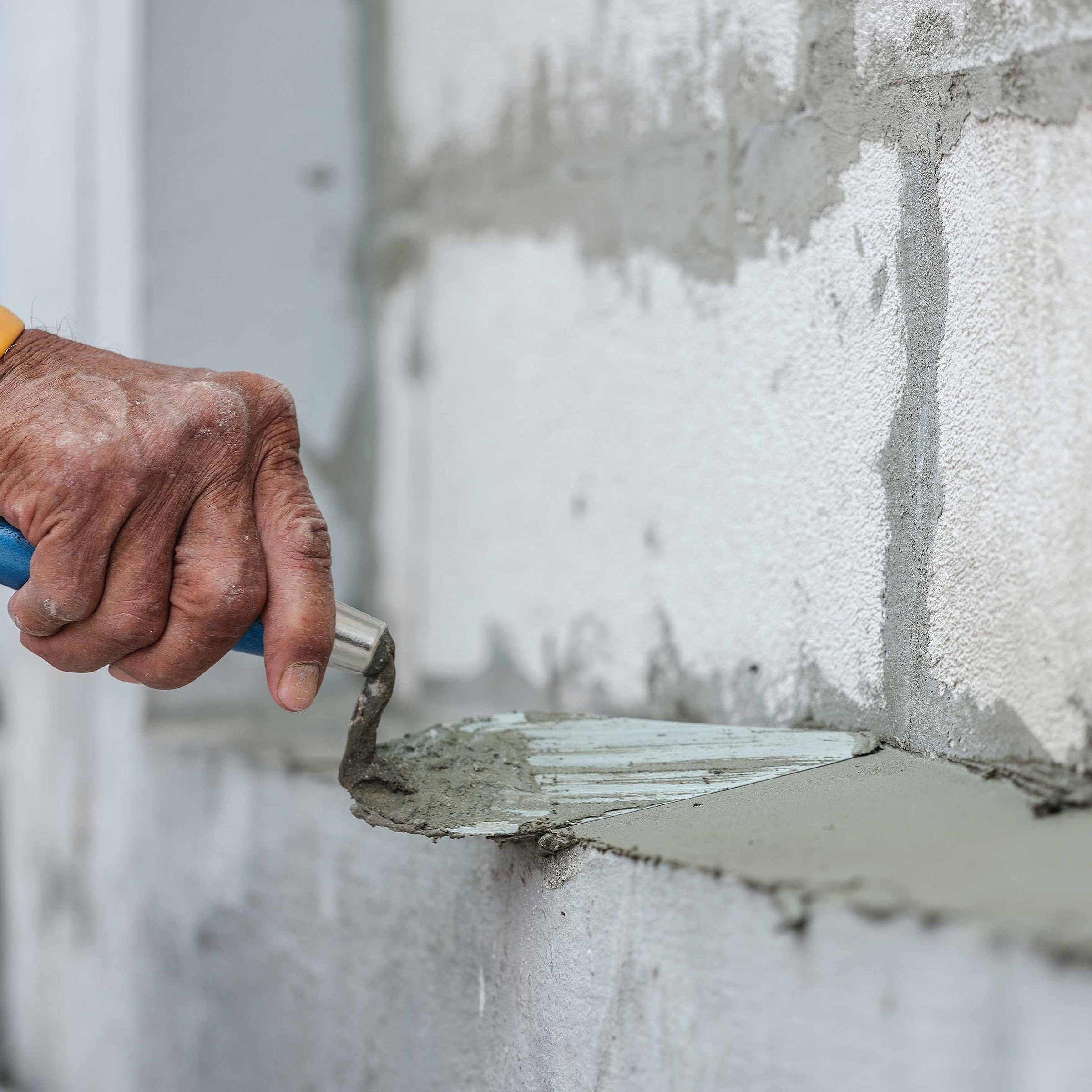A hand is applying fresh mortar onto a brick using a trowel