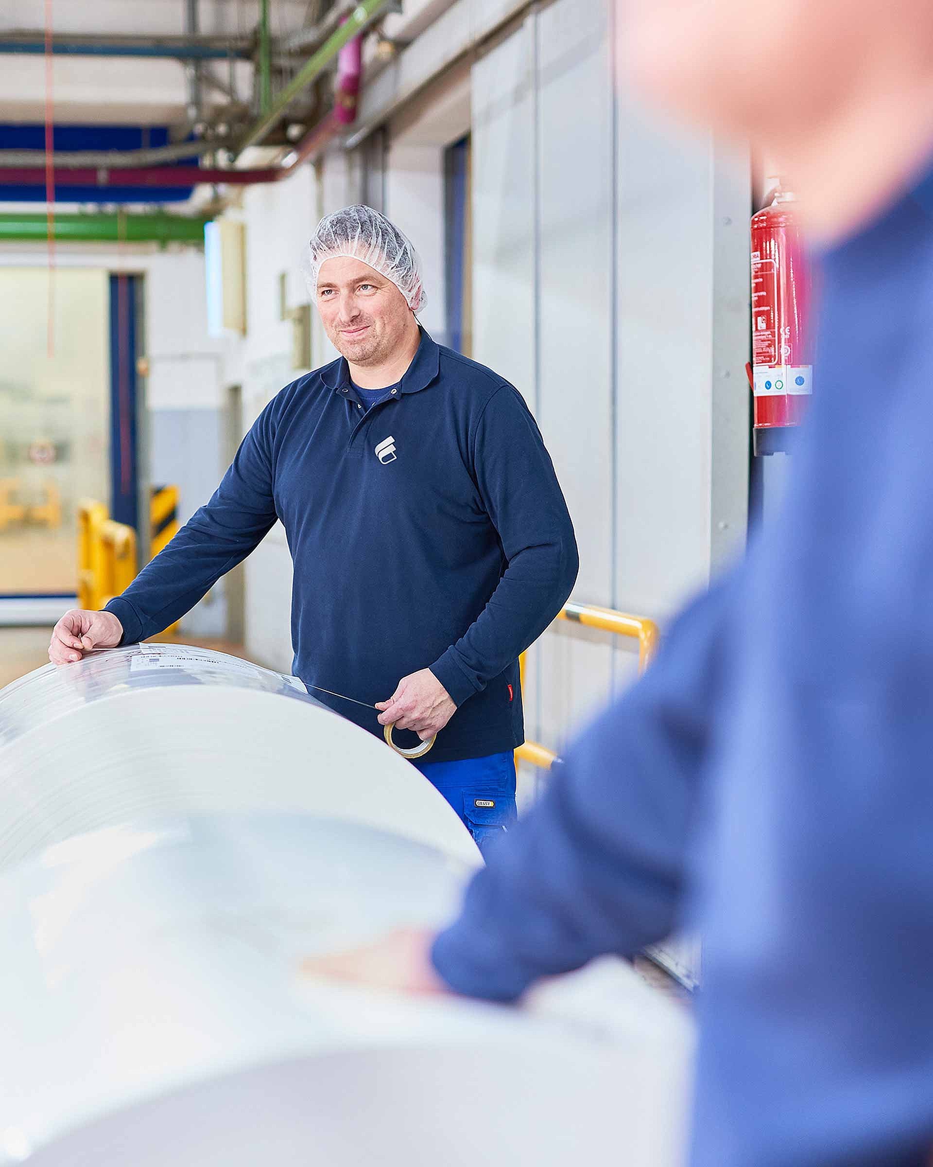 An employee wearing Bischof+Klein clothing and a hair net is standing behind a large reel of plastic film in the production department