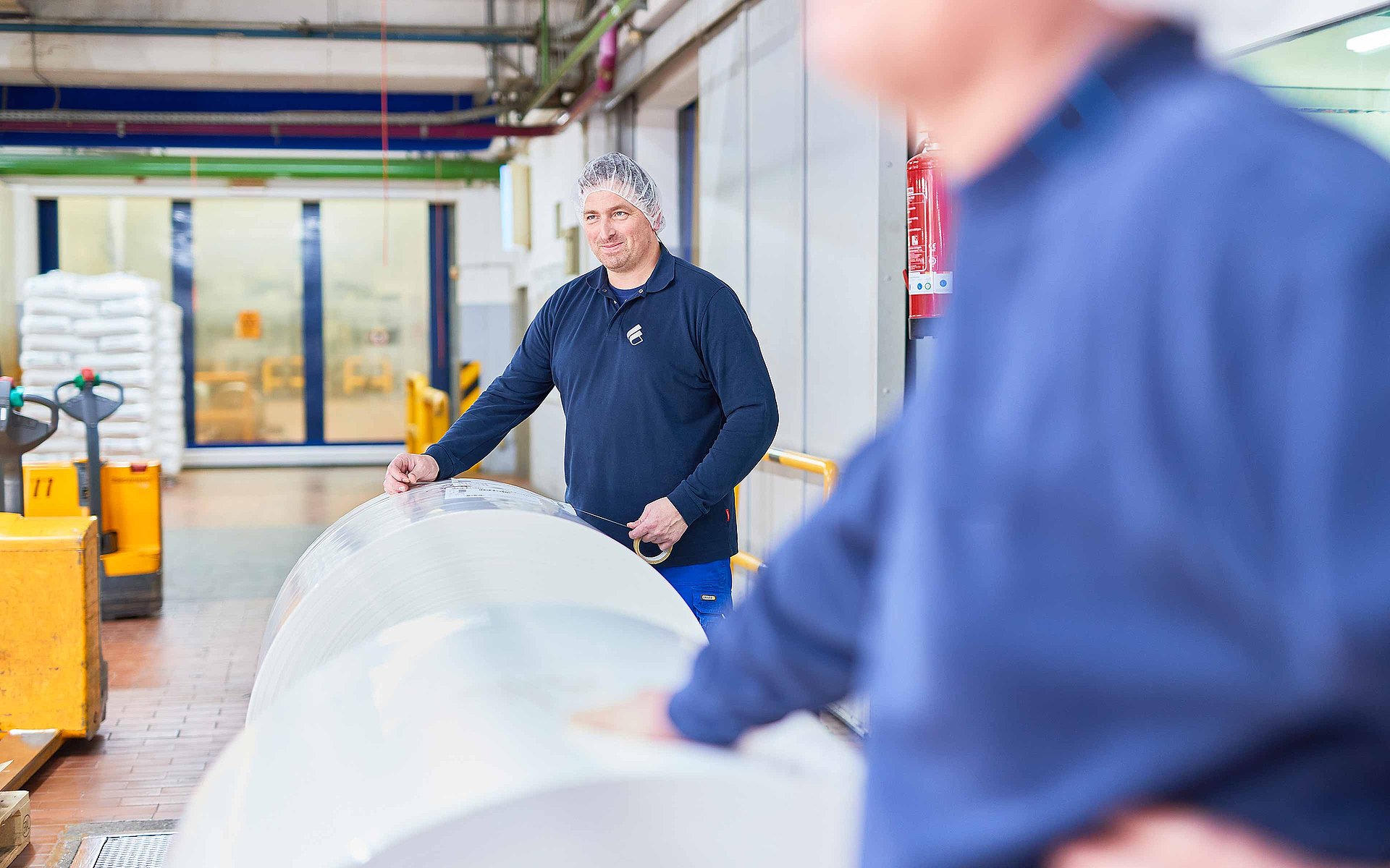 Un collaborateur en vêtement de travail de Bischof+Klein et portant une résille se tient à la production derrière une grosse bobine de film plastique