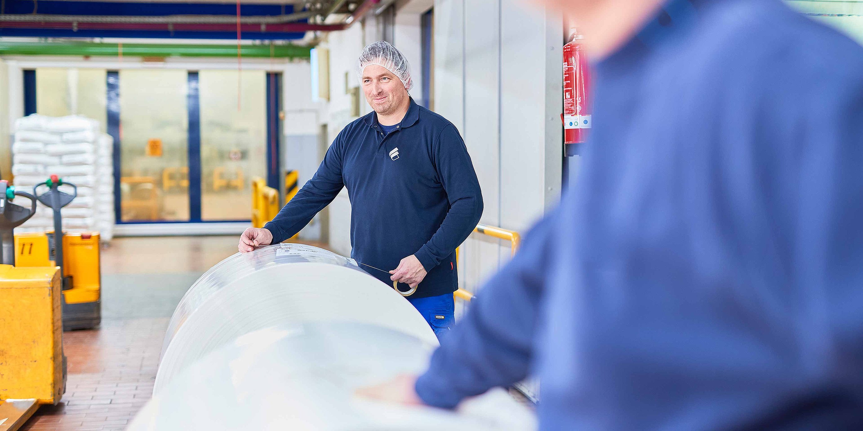 An employee wearing Bischof+Klein clothing and a hair net is standing behind a large reel of plastic film in the production department
