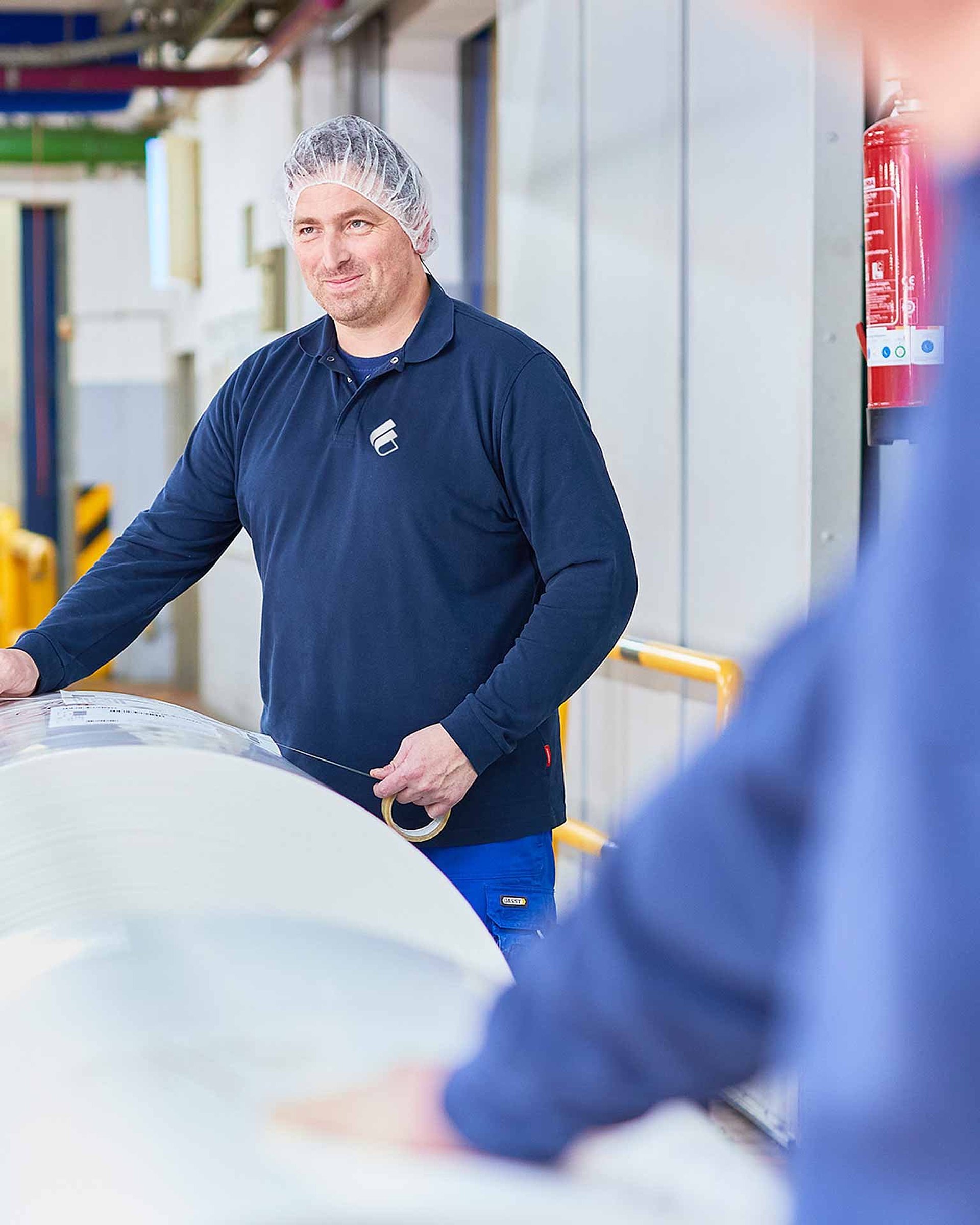 Ein Mitarbeiter mit Bischof+Klein Kleidung sowie Haarnetz steht in der Produktion hinter einer großen Rolle mit Kunststofffolie