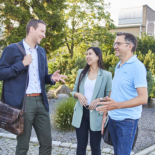 Drei Mitarbeitende stehen in Business Kleidung vor dem Bürogebäude und unterhalten sich lächelnd