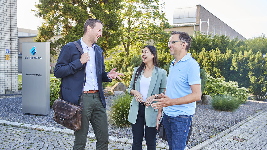 Drei Mitarbeitende stehen in Business Kleidung vor dem Bürogebäude und unterhalten sich lächelnd