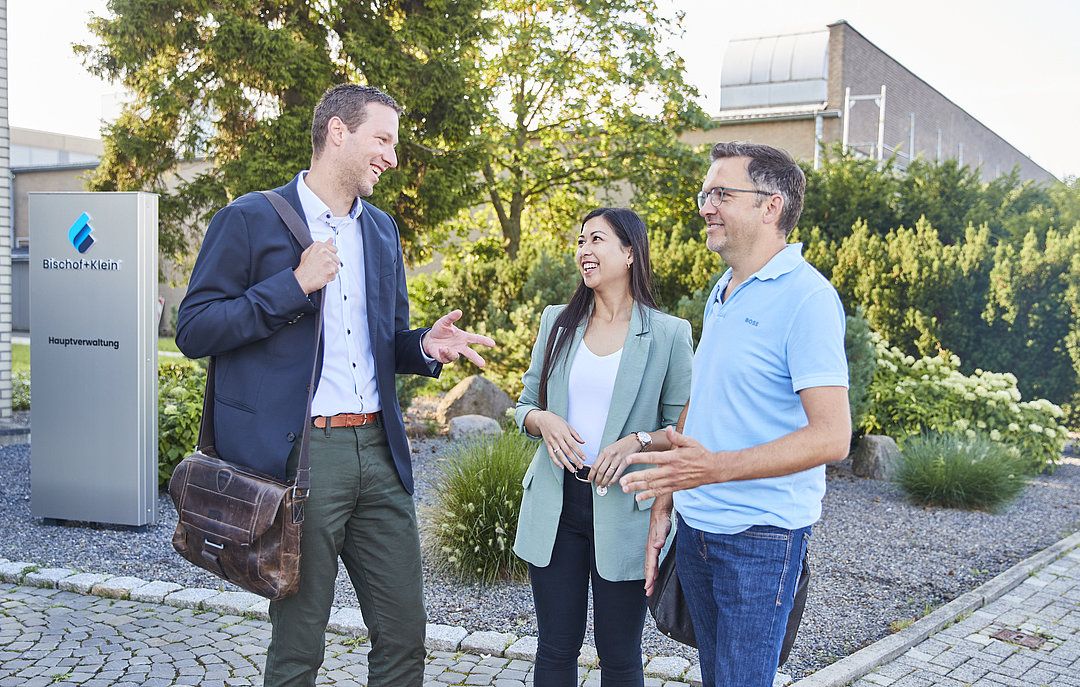 Drei Mitarbeitende stehen in Business Kleidung vor dem Bürogebäude und unterhalten sich lächelnd